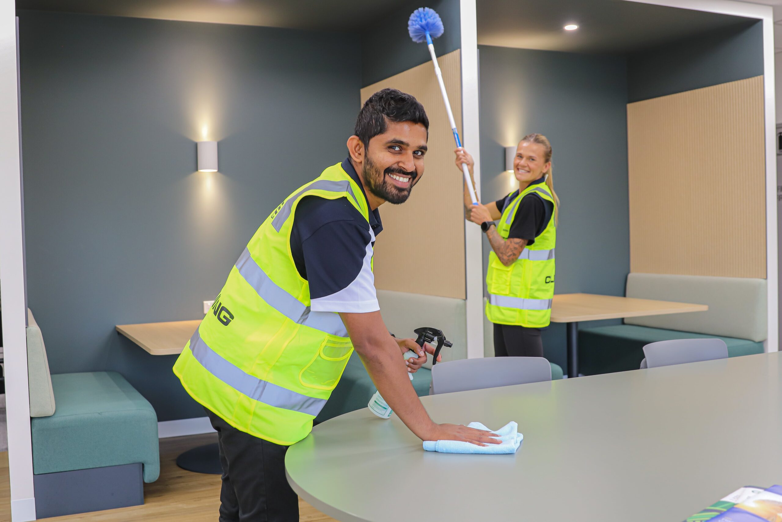In a bright office space, two people wearing high-visibility vests focus on office cleaning tasks. One wipes a table, while the other carefully cleans a ceiling corner with a brush, ensuring every detail in this modern workspace is spotless.