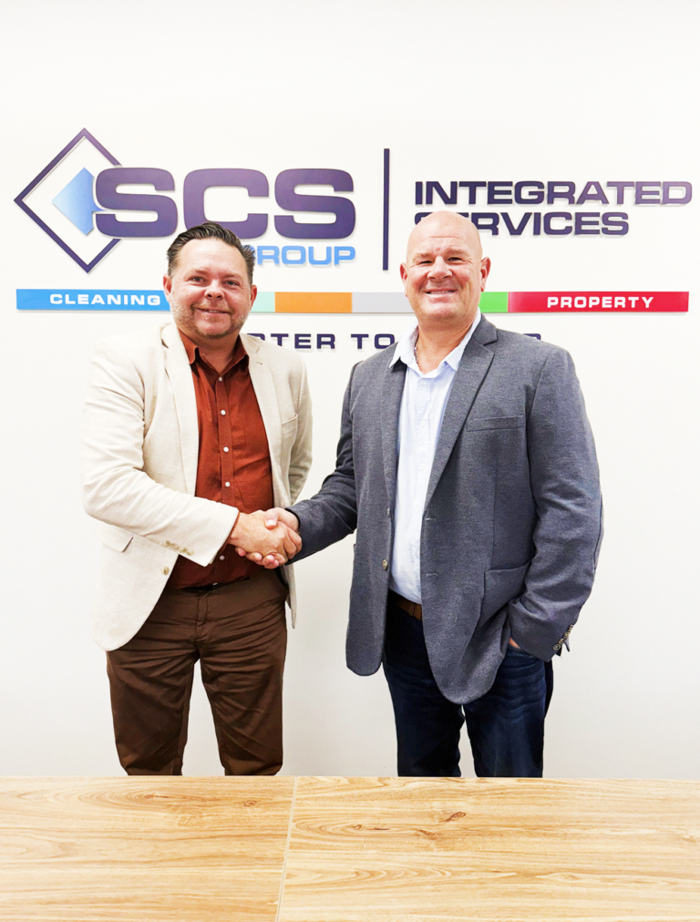 SCS Group Integrated Services Co-Founders Scott McCartney and Nicholas Pastras shaking hands in front of the company's branded wall featuring coloured service category banners including Cleaning and Property, with a light timber table in the foreground. Scott McCartney wears a cream blazer over a rust-coloured shirt, and Nicholas Pastras wears a grey blazer over a light blue shirt.