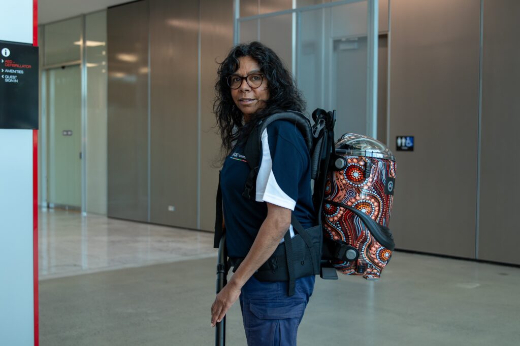 An SCS Indigenous female cleaning team member wearing a navy and white branded polo shirt and glasses, holding a mop handle while carrying a backpack vacuum cleaner decorated with Aboriginal dot art in warm tones of red, orange, and blue, standing in a modern commercial building corridor.