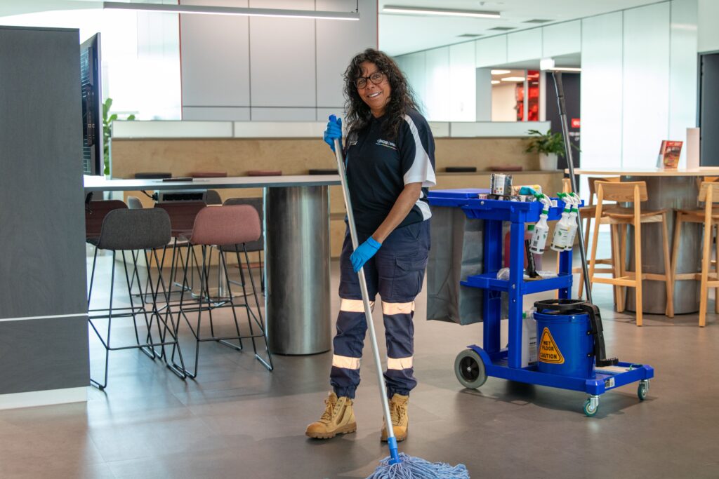 Professional commercial cleaner mopping the floor in a modern office break area, with a fully stocked cleaning trolley, tables, chairs, and kitchen facilities in the background.