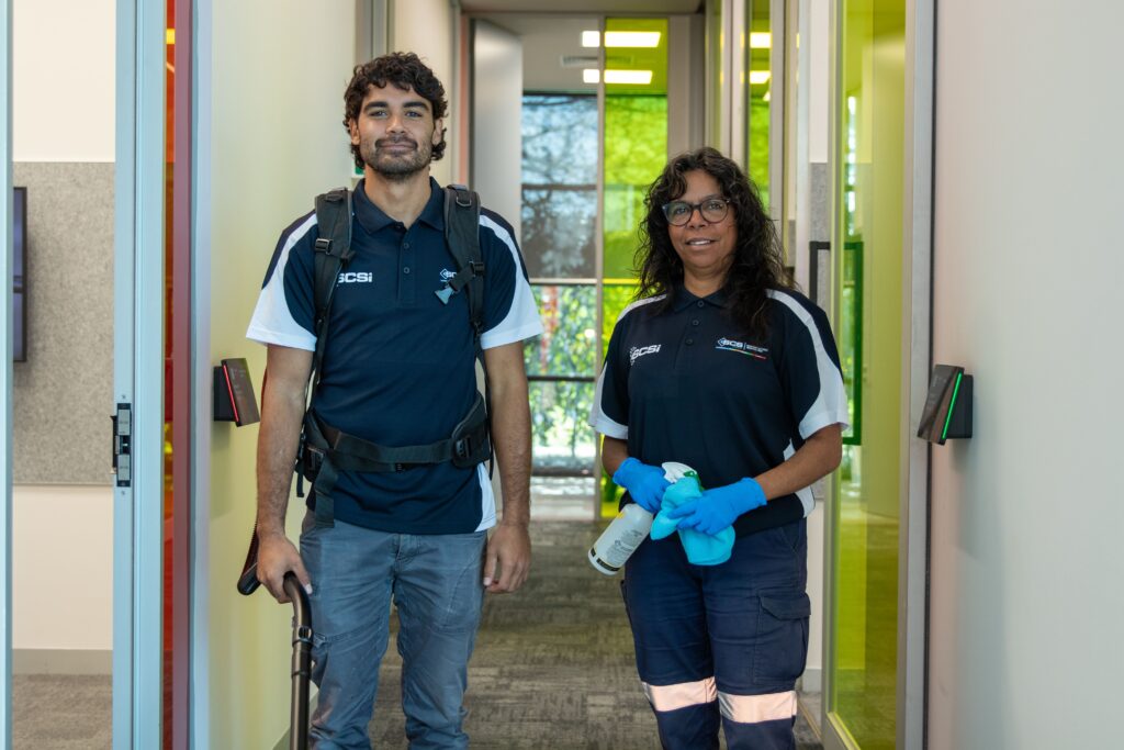 Two professional commercial cleaning staff standing in a modern office corridor, with one holding disinfectant spray bottles and the other carrying a vacuum cleaner backpack and hose, ready to provide cleaning services.