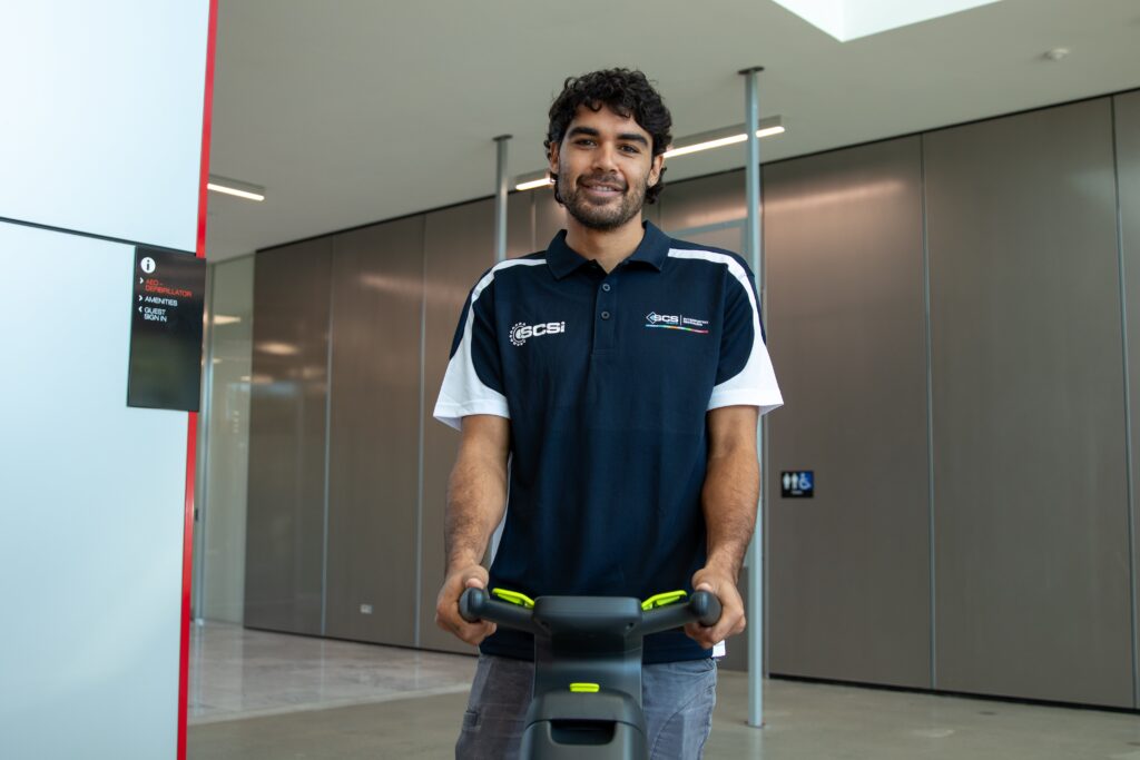 Professional commercial cleaner operating a floor cleaning machine in a modern office building lobby, wearing an SCS uniform and preparing to clean hard floor surfaces.