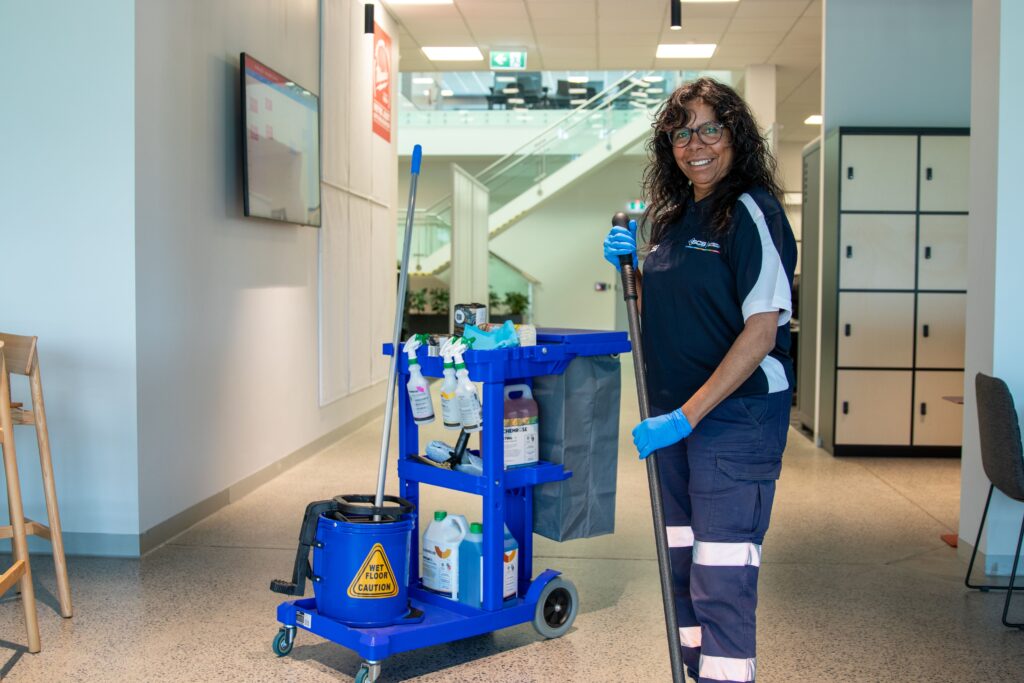 Professional commercial cleaner standing beside a fully stocked cleaning trolley in a modern office hallway, holding a mop and wearing protective gloves while preparing to clean the workplace.