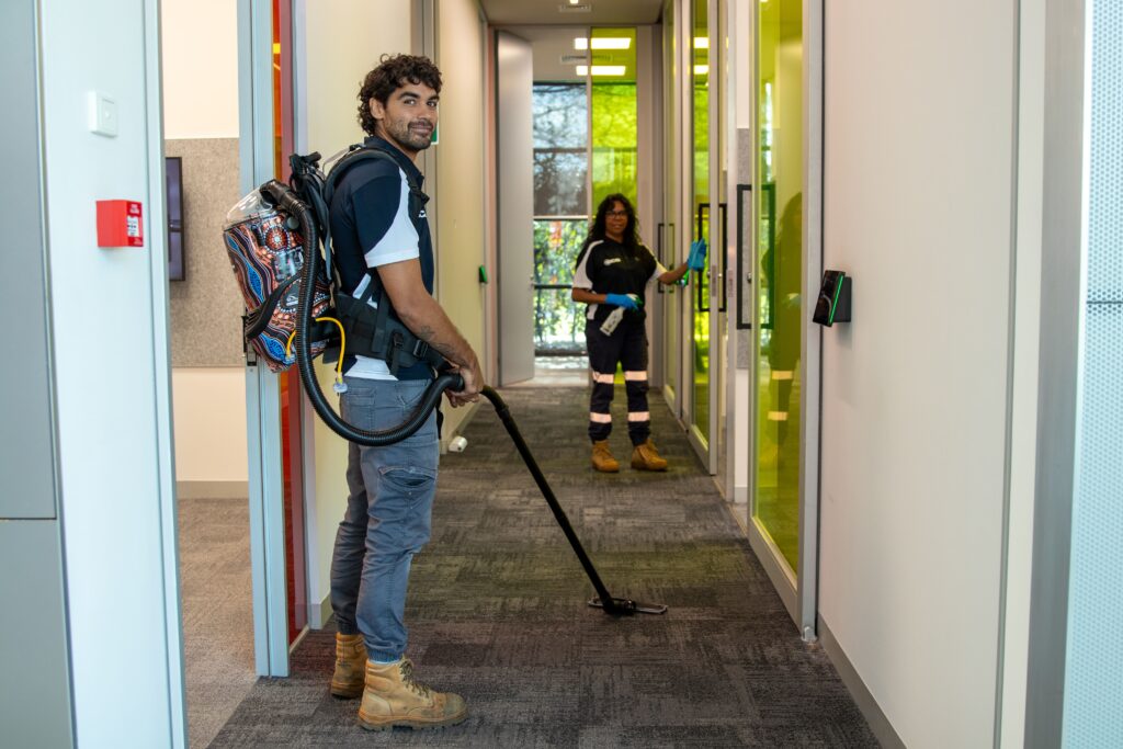 Professional commercial cleaner using a backpack vacuum to clean a modern office corridor while a colleague disinfects glass doors in the background, demonstrating a coordinated workplace cleaning service.