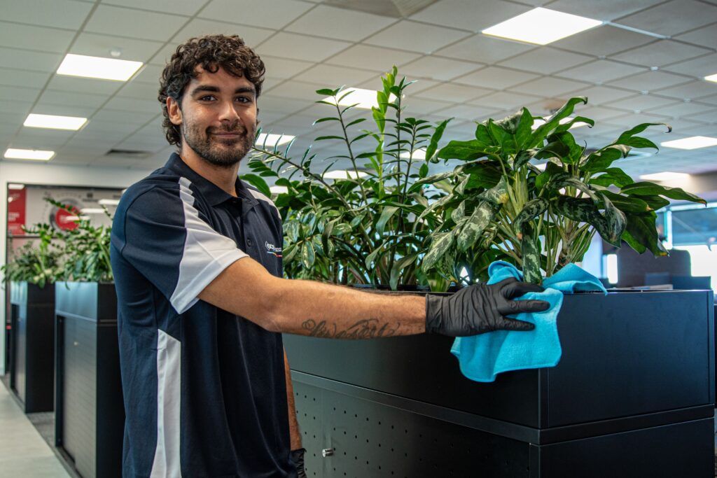 An SCS Indigenous male cleaning team member smiling at the camera while wiping down a dark office cabinet with a blue microfibre cloth and black gloves, wearing a navy and white branded polo shirt, with a row of lush indoor plants and a modern open-plan office visible in the background.