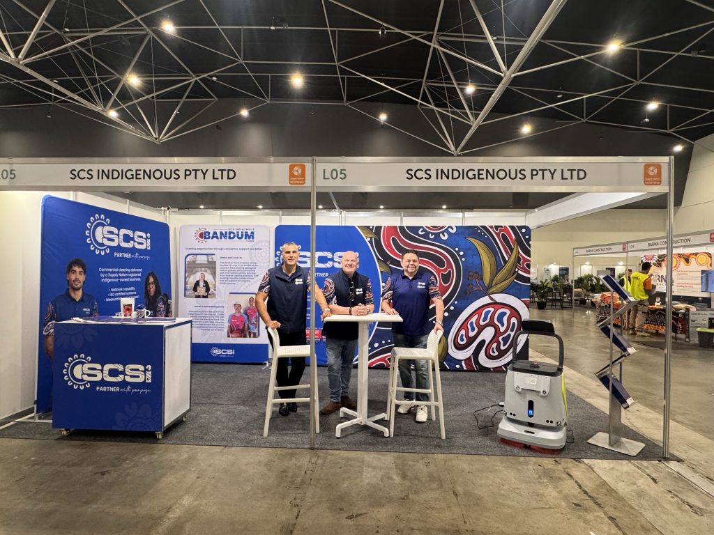 SCS Indigenous team at trade show booth L05 with SCSi banners, Aboriginal artwork backdrop, and a robotic floor cleaner.