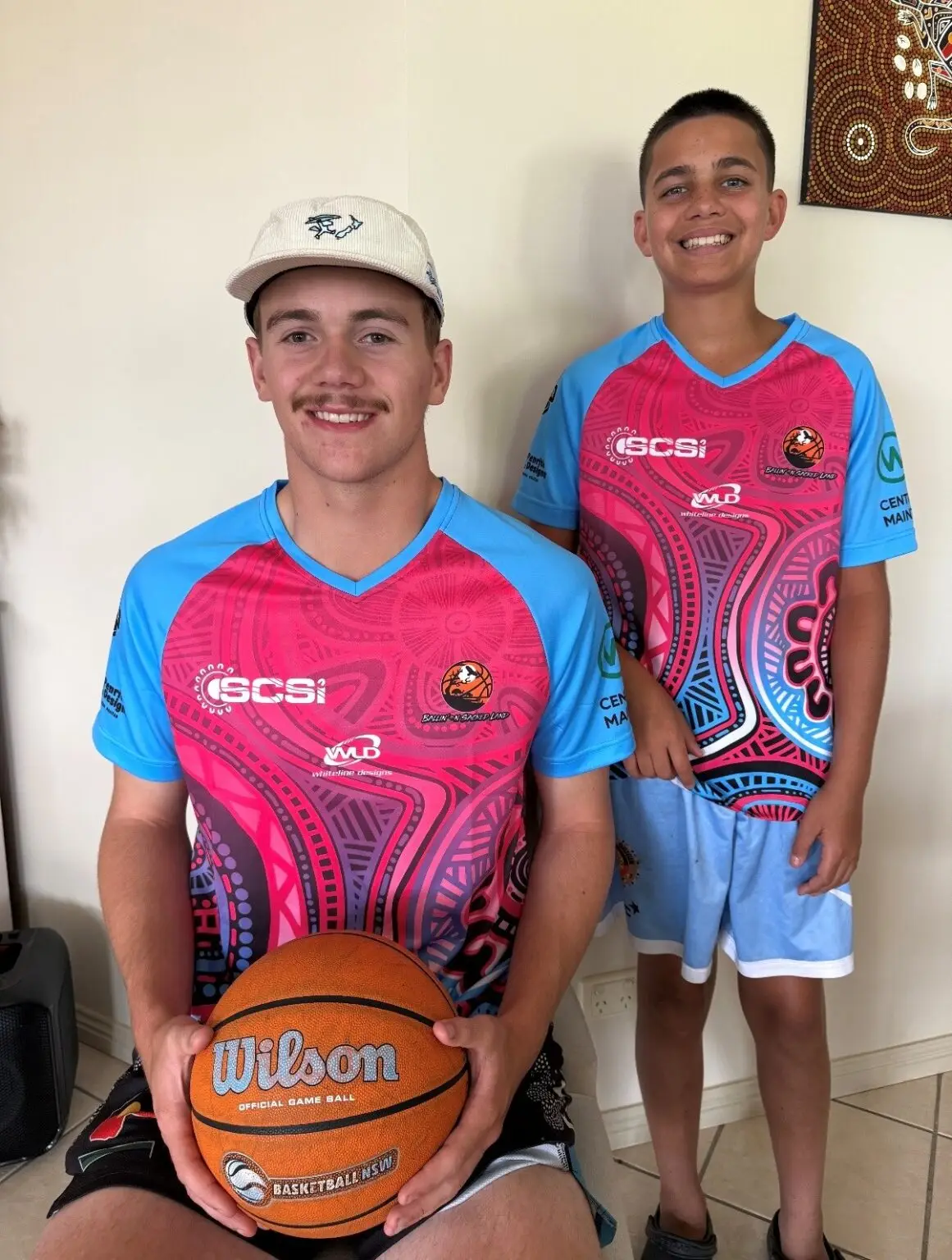 Hugo and Jurrah wear matching pink, blue, and purple Indigenous-print SCSI basketball jerseys. Hugo sits in front holding a Wilson basketball, and Jurrah stands behind him. An Aboriginal dot painting hangs on the wall in the background