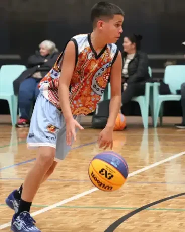 A young basketball player named Jurrah dribbles the ball on an indoor court, wearing an Indigenous-print jersey with orange, red, and white patterns, light blue shorts, and dark sneakers, with spectators seated in the background.