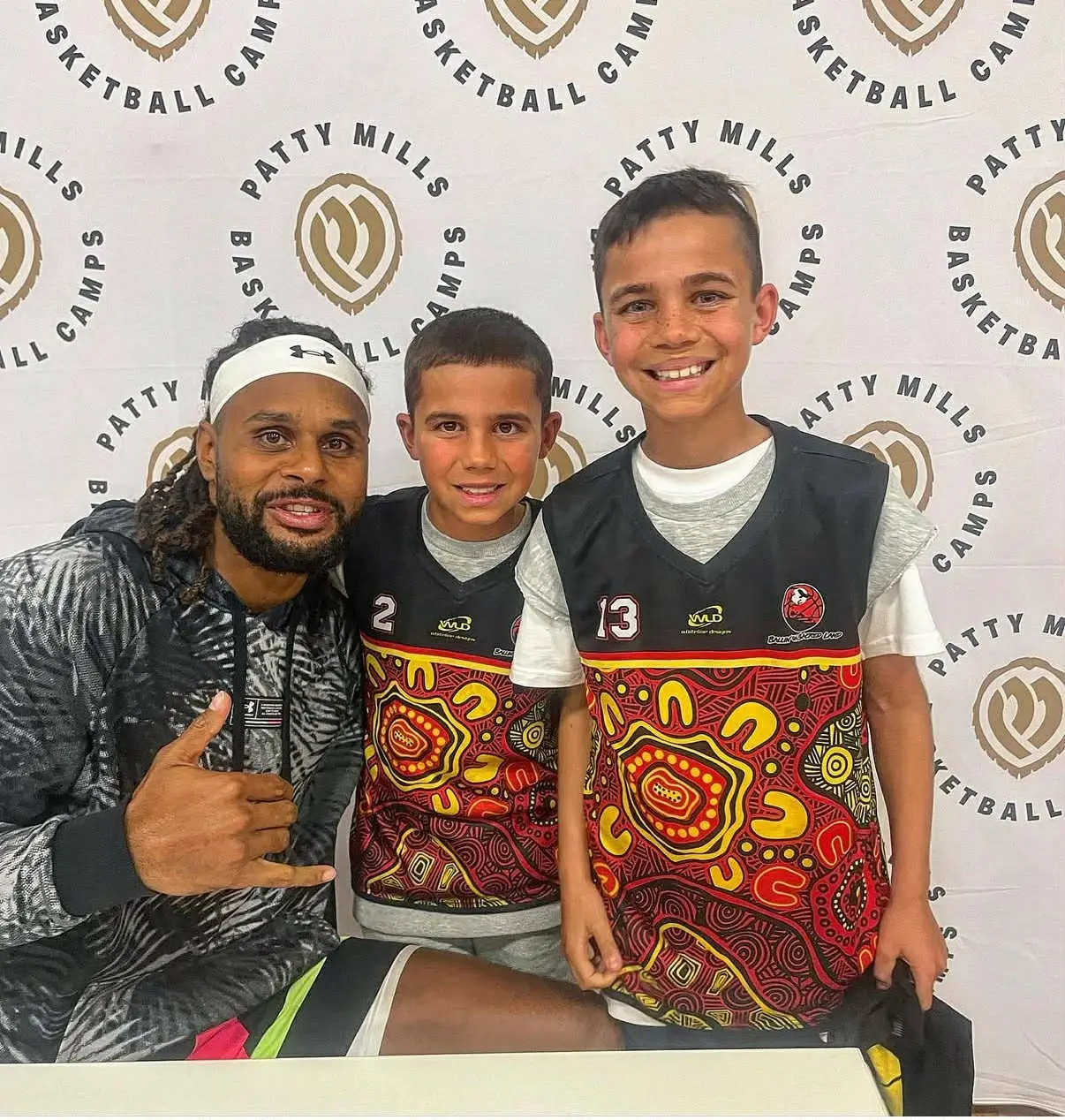 NBA player Patty Mills poses with two young basketball players, Jurrah and a teammate, in front of a Patty Mills Basketball Camps step and repeat backdrop. Mills wears a headband, patterned jacket, and shorts, and is making a shaka hand gesture. The two boys wear matching black jerseys with red, yellow, and black Indigenous-print designs, numbered 2 and 13