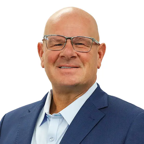 Professional portrait of Nicholas Pastras, smiling, wearing glasses and a navy suit jacket with a light blue shirt, against a white background.