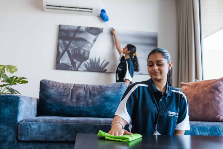 Two SCS cleaning staff members working in a modern living room, one wiping a coffee table with a microfibre cloth and the other dusting a wall-mounted air conditioning unit.