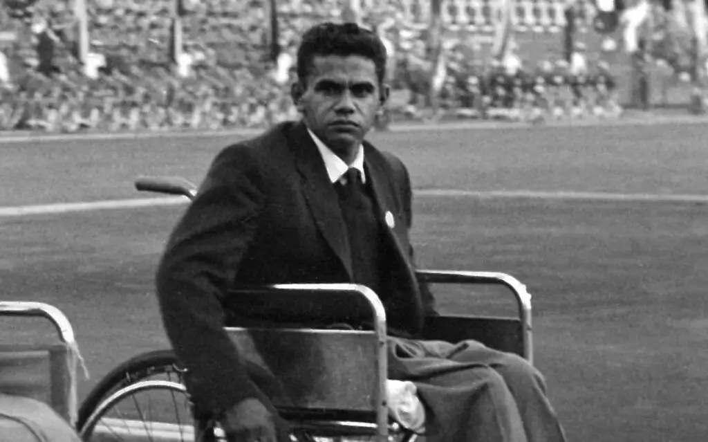 Black and white archival photo of Uncle Kevin Coombs seated in a wheelchair on a sports field, wearing a suit and tie, looking directly at the camera, with a stadium crowd visible in the background