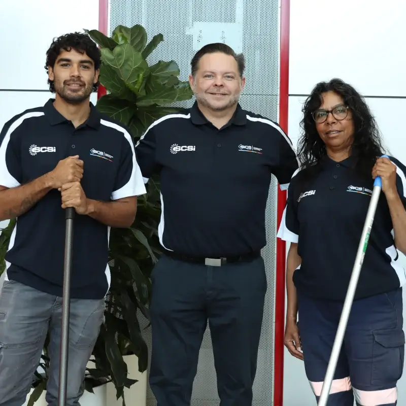 Three SCS Indigenous team members in navy and white branded polo shirts posing in a modern commercial building, with a large indoor plant and red architectural feature wall behind them. The two outer team members hold mop handles, while the team member in the centre stands between them smiling.